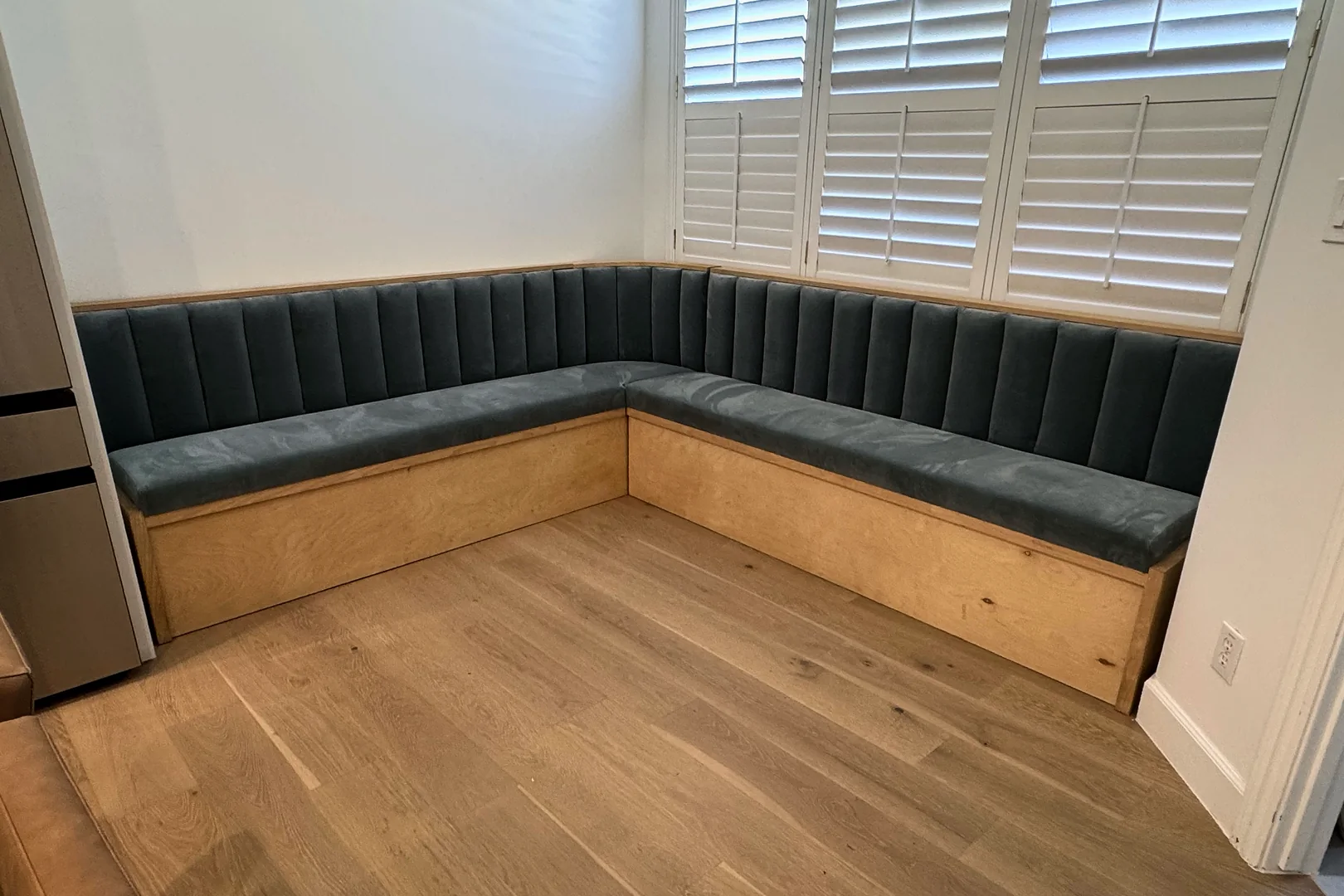 L-shaped corner banquette with a vertically channelled back and a blue-grey upholstered seat, on a plywood storage base beside a window with white shutters