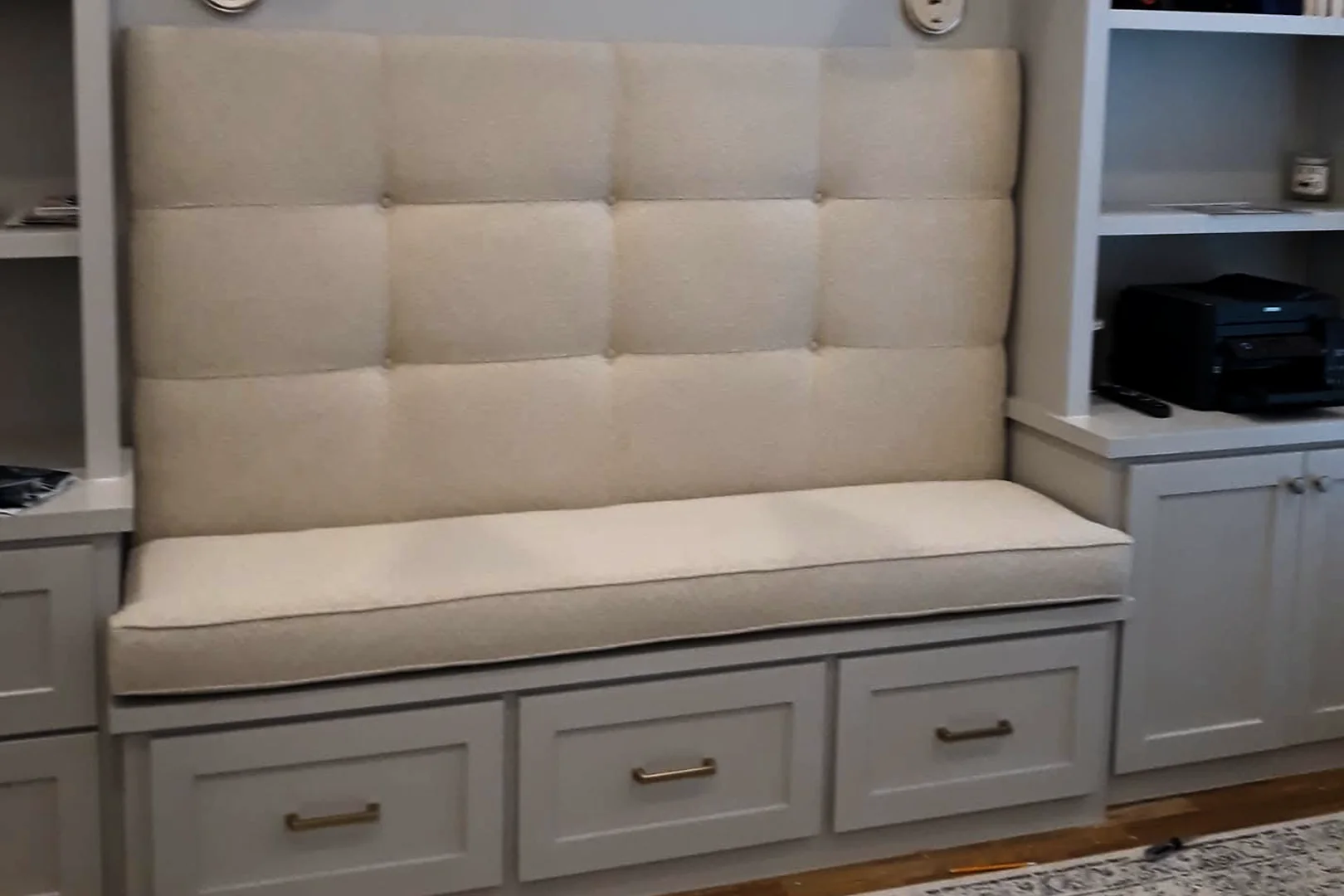 Built-in window seat with a tall, rectangular-tufted cream back and a cream seat cushion above three drawers, between white shelves