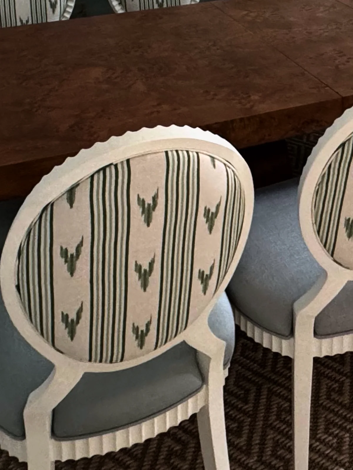 Close-up of oval chair backs covered in a green and cream striped fabric with a leaf motif, set within white fluted frames, around a wood table