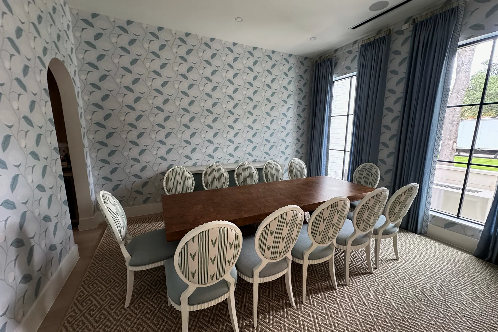 A dining room with a long wood table and a row of white oval-back chairs with striped backs and pale blue seats, against leaf-patterned wallpaper