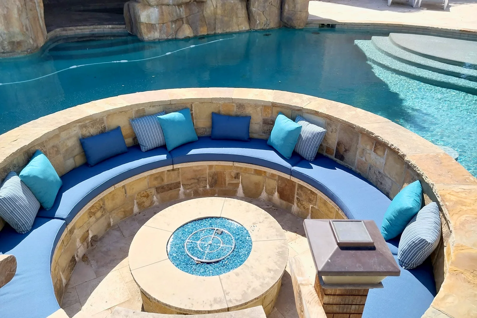 Circular stone seat around a fire pit beside a swimming pool, with blue cushions and blue, aqua and striped pillows