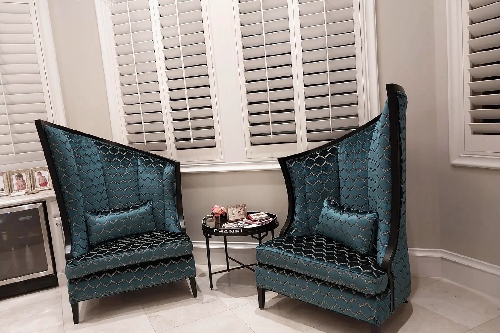 Two tall wing chairs with dark wood frames, upholstered in teal patterned velvet with matching lumbar pillows, in front of a window with white shutters